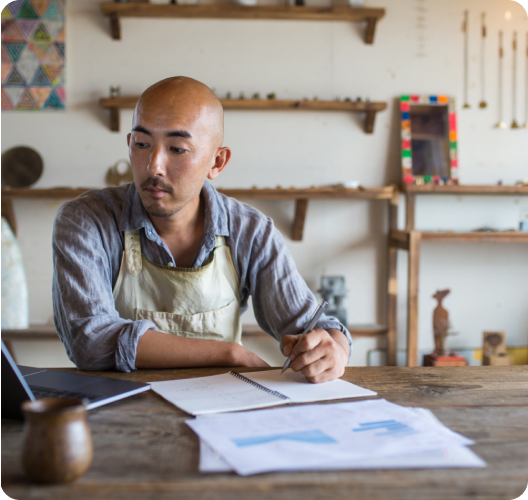 A man wearing a work apron sits at a table. His laptop is open and he’s holding a pen, taking notes.