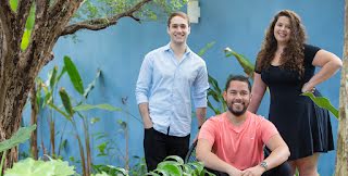 Cuidas founders João, Matheus, and Deborah pose together in front of a blue wall.