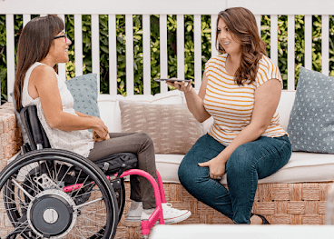 Eine Frau sitzt in einem Rollstuhl auf einer Terrasse und unterhält sich mit einer Freundin, die ein Android-Smartphone in der Hand hält.