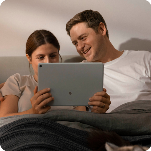 Deux personnes se détendent dans un lit et sourient en regardant un divertissement sur leur tablette.