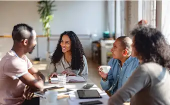 Four team members sit at a table having a coffee-work meeting with notebooks, tablets and phones across the table