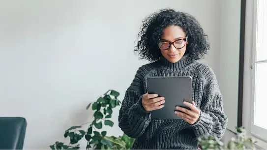 Una giovane donna con un maglione grigio guarda lo schermo di un tablet. È in piedi accanto a una finestra illuminata dal sole e dietro di lei si vede una pianta.