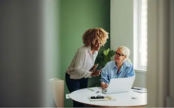 Dos mujeres se reúnen frente a la pantalla de una laptop y hablan sobre el trabajo.