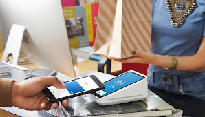 Shopper purchasing with touchless mobile checkout, woman cashier in blue t-shirt hands over diagonally striped paper shopping bag.