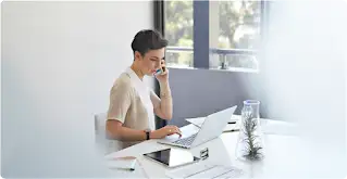 Une femme aux cheveux courts est assise à son bureau, sur lequel sont posés un ordinateur portable et une tablette. Elle parle au téléphone avec un smartphone.