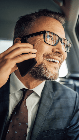 A white man wearing a gray checkered suit and burgundy tie talking on his mobile phone while looking out the window in the backseat of a car.