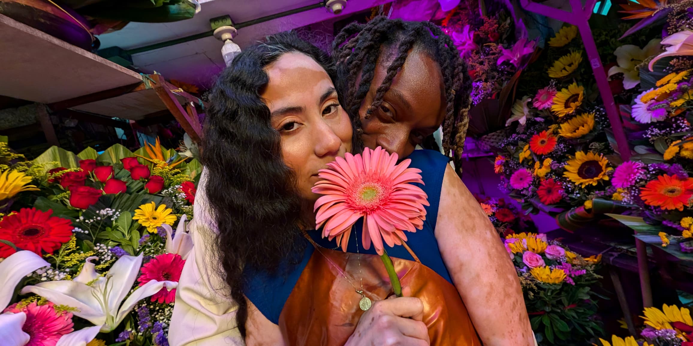 Two people pose facing the Pixel Camera, surrounded by flowers. Colors and skin tones are vividly captured by Pixel 9.