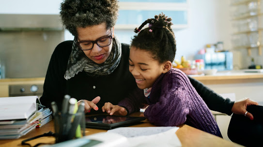 A mother and daughter sit at their kitchen table, interacting with a tablet device. The daughter smiles.