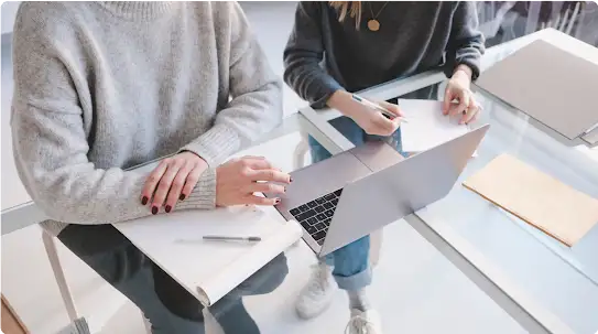 Due persone lavorano sedute una accanto all’altra a una scrivania dal piano in vetro su cui si vede un laptop.