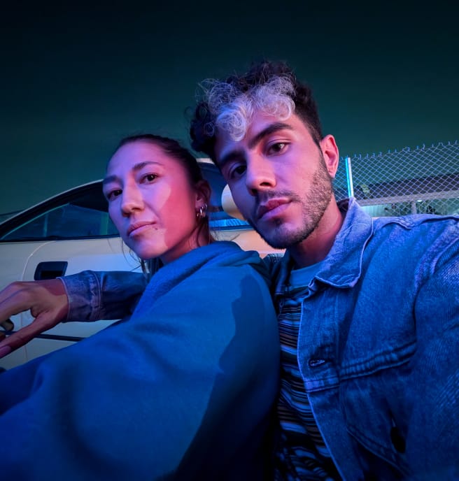 Two people pose in dim blue light., angled away but looking at the camera