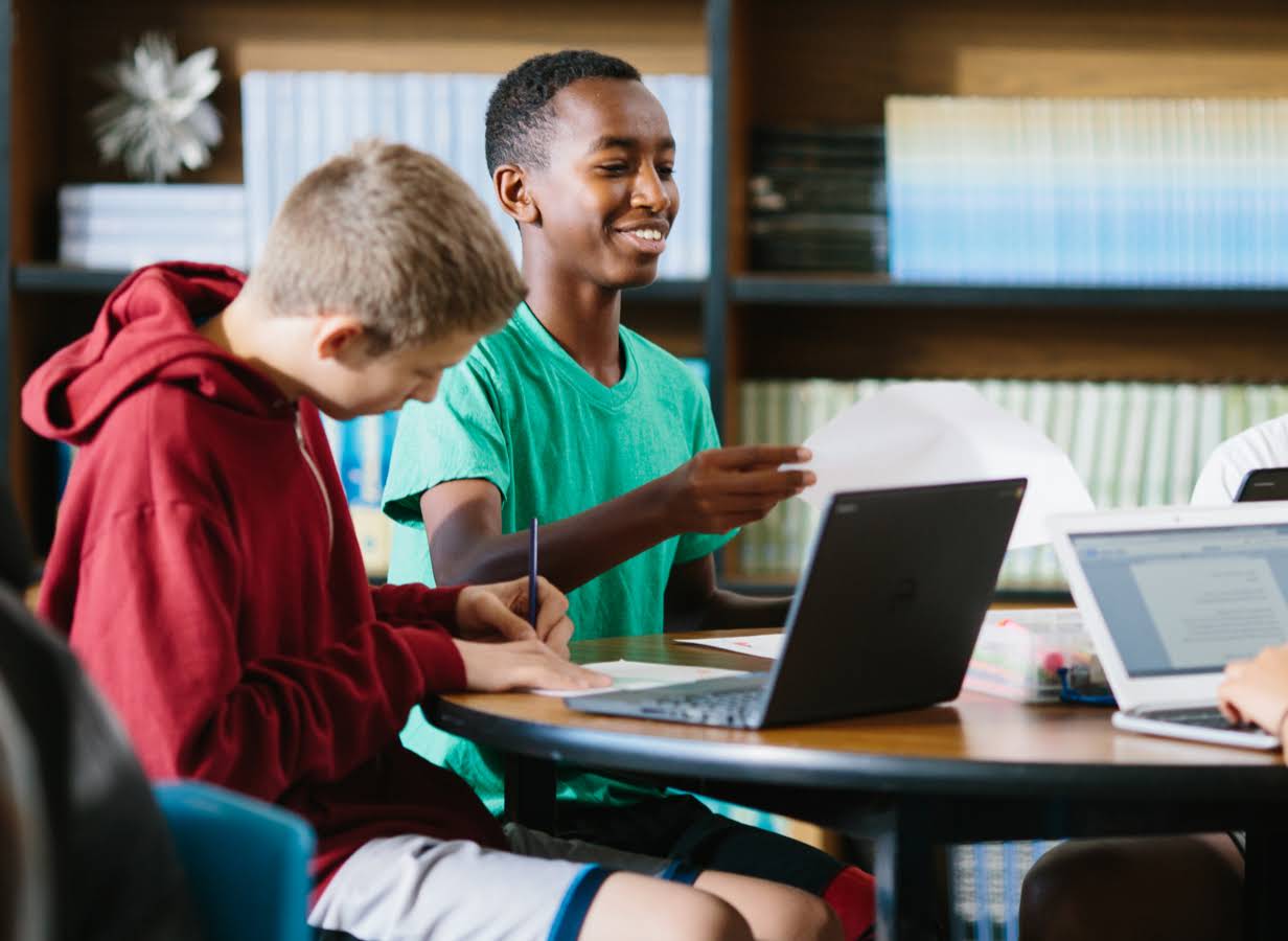 Un alumno sujeta una hoja de papel cerca de un compañero que trabaja frente a una Chromebook.