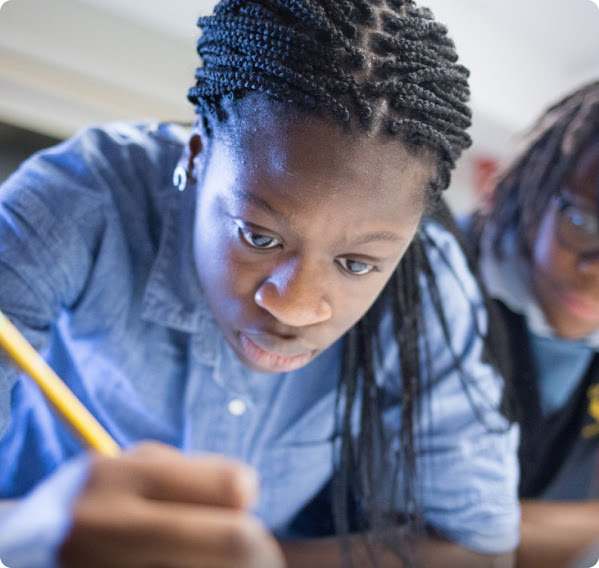 A young girl sits at a desk with a pencil in hand, working hard. Another girl in the background looks on.