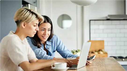 Due donne usano un laptop. Una, con una maglia bianca, digita e l’altra, con una maglia blu, tiene in mano uno smartphone e guarda lo schermo del laptop.