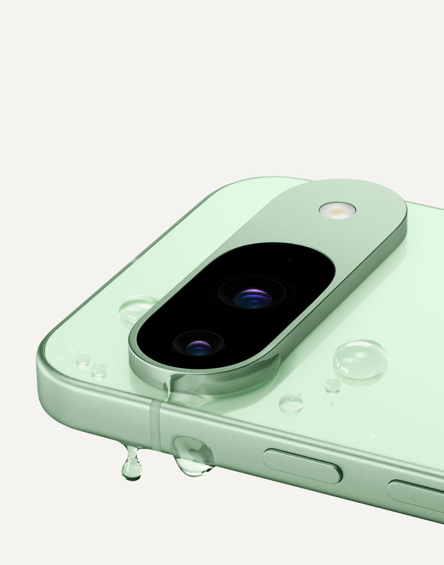 A close up of the top corner of Pixel 9 in Wintergreen color, one of its camera lenses in view. A single drop of water drips off the side of the phone.