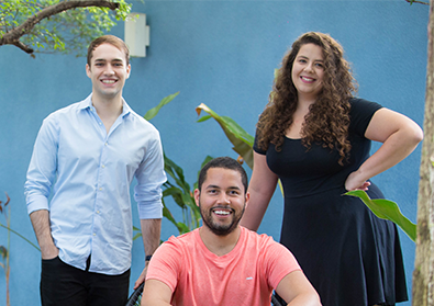 Cuidas founders João, Matheus, and Deborah pose together in front of a blue wall.