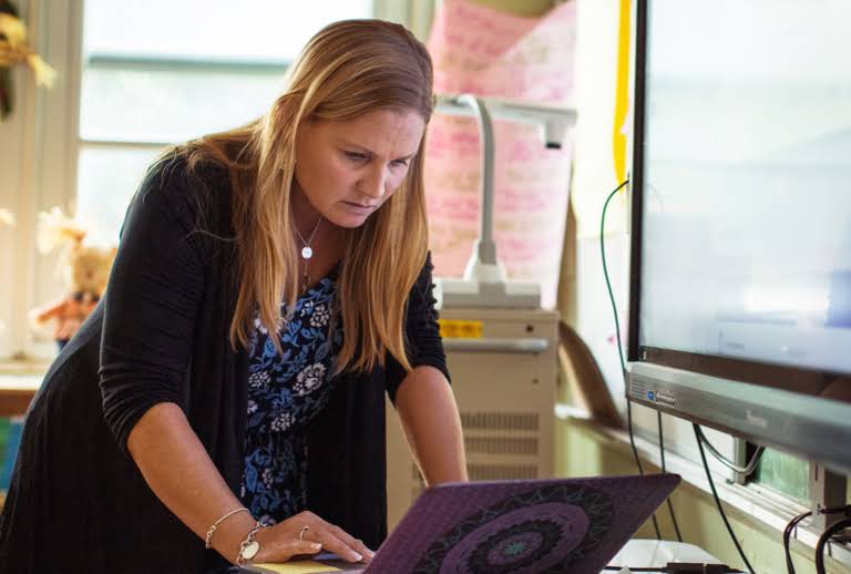 Una educadora se inclina sobre una laptop que está apoyada en una mesa dentro de un aula.