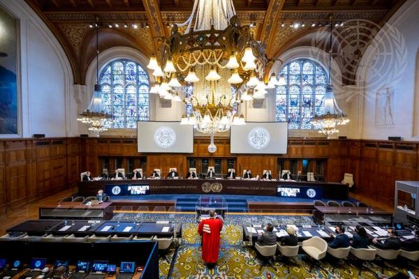 A wide view of the courtroom with justices seated at the head of the room. A man in a red robe stands before a podium at the center facing the justices.