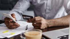 A man seated at a desk covered in papers and bills holds a phone in one hand and a credit card in the other