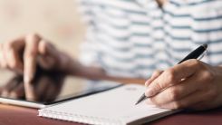 A person writing in a notebook and referencing an iPad