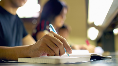 A close-up photograph of a student's hand taking notes in a notebook. They are in a classroom setting.