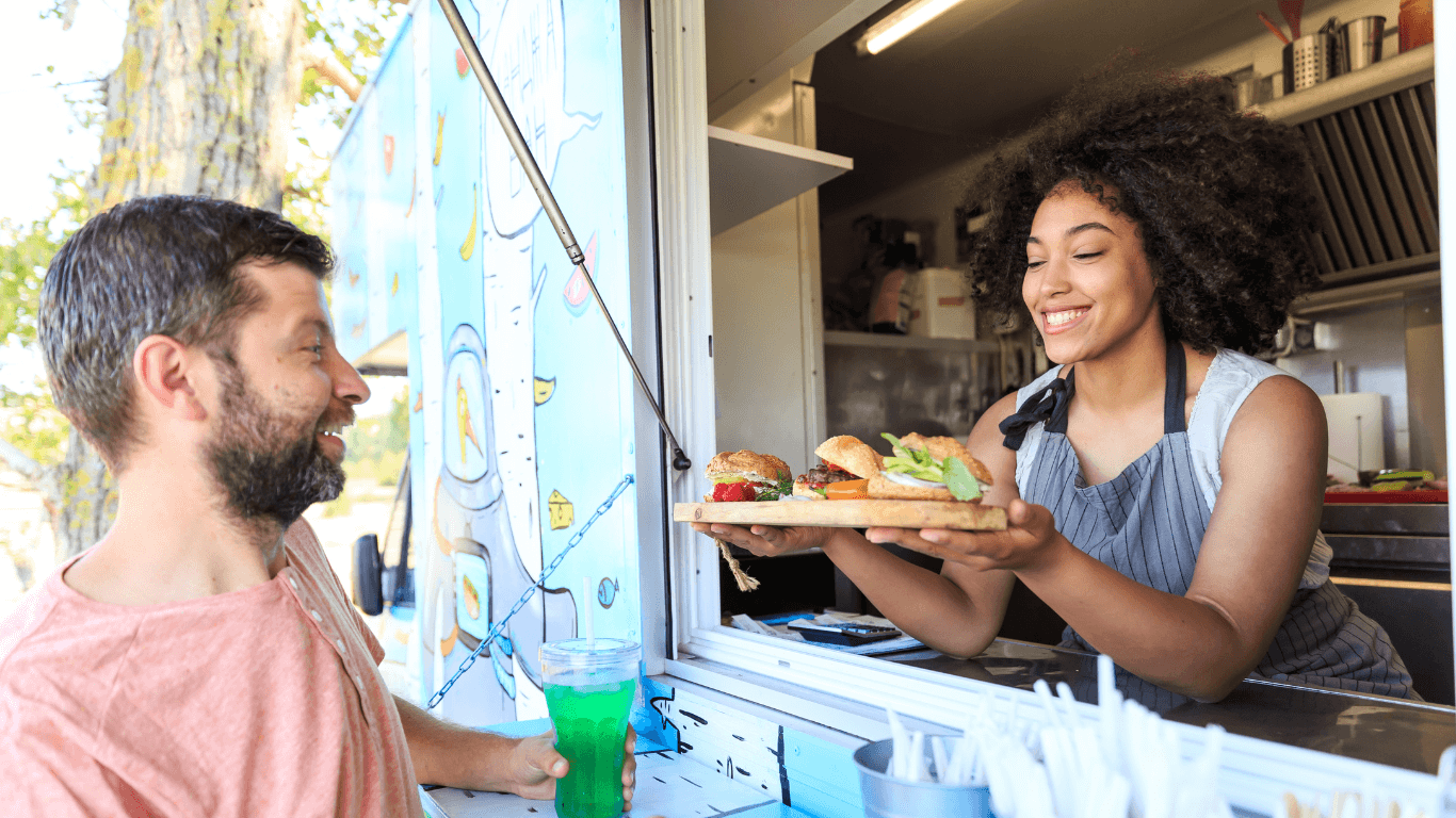 Smiling woman serving a variety of sandwiches to a man from a food truck window