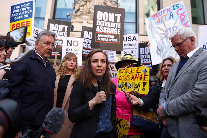 A woman with long dark hair speaks into a microphone in front of a crowd of protesters holding signs.