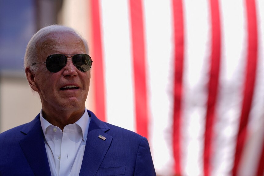 A man with grey hair wearing sunglasses, dressed in a white shirt and blue blazer with a US flag pin 