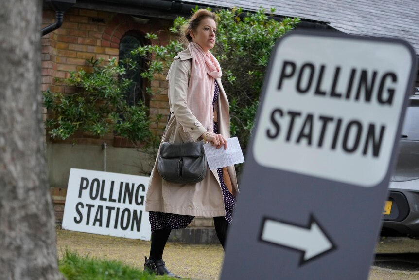 A woman holds a document as walks on pavement past signs saying "polling station".