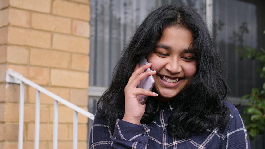 Girl with black hair standing outside, talking on her phone, smiling.