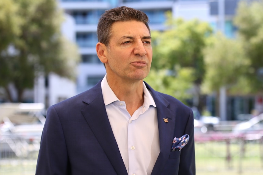 Basil Zempilas speaking into microphones outdoors on a sunny day, wearing a dark suit and white shirt.