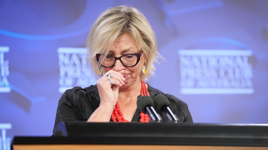 A white woman, trying not to cry. She's standing at a lectern in front of a purple background.