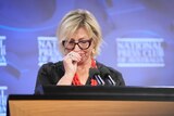 A white woman, trying not to cry. She's standing at a lectern in front of a purple background.