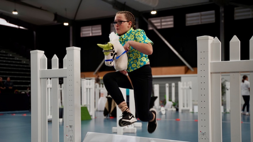 A young girl leaps over a static jump while riding a hobby horse