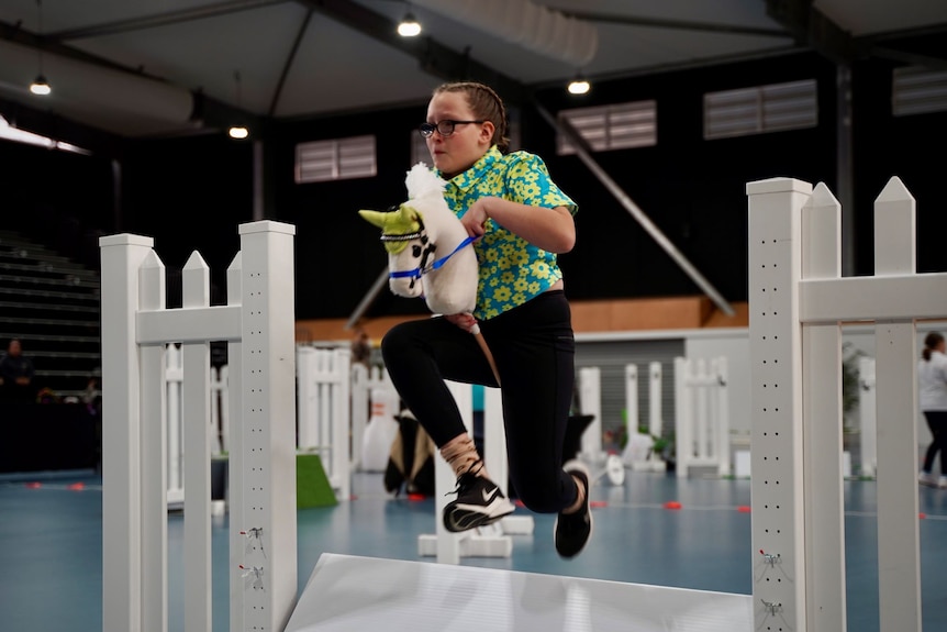 A young girl leaps over a static jump while riding a hobby horse