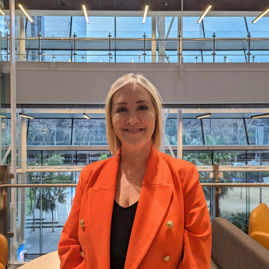 A blonde woman in an orange jacket smiles at the camera