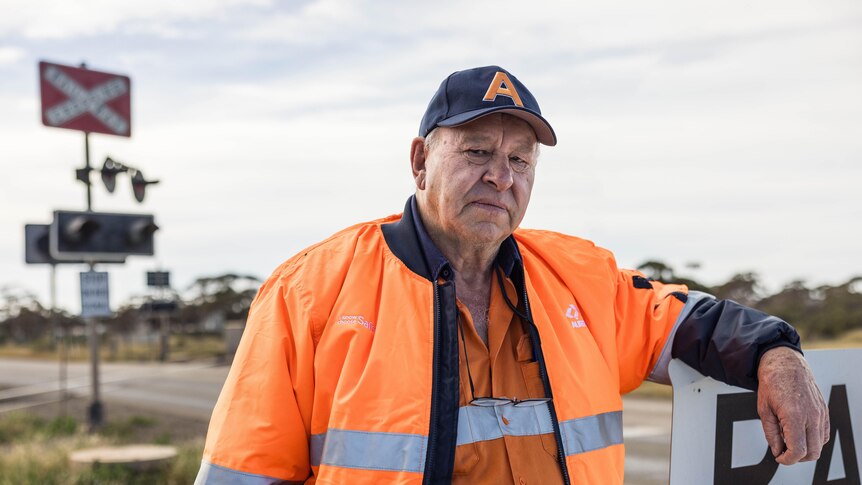 A man wearing high-vis orange workwear leaning on a railway sign.  