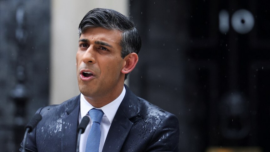 Rishi Sunak stands in a blue suit speaking behind a lectern in the rain in the street