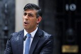 Rishi Sunak stands in a blue suit speaking behind a lectern in the rain in the street