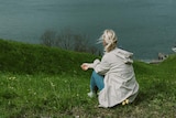 A woman with a blonde pony sits on a grassy knoll looking away from the camera to the ocean below.