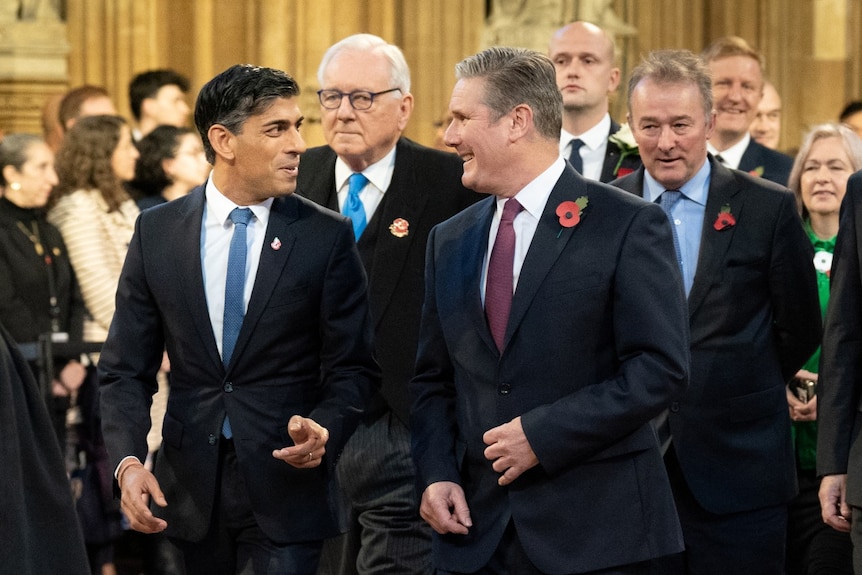 Two men in suits smiling at each other, with lines of other men behind them.,