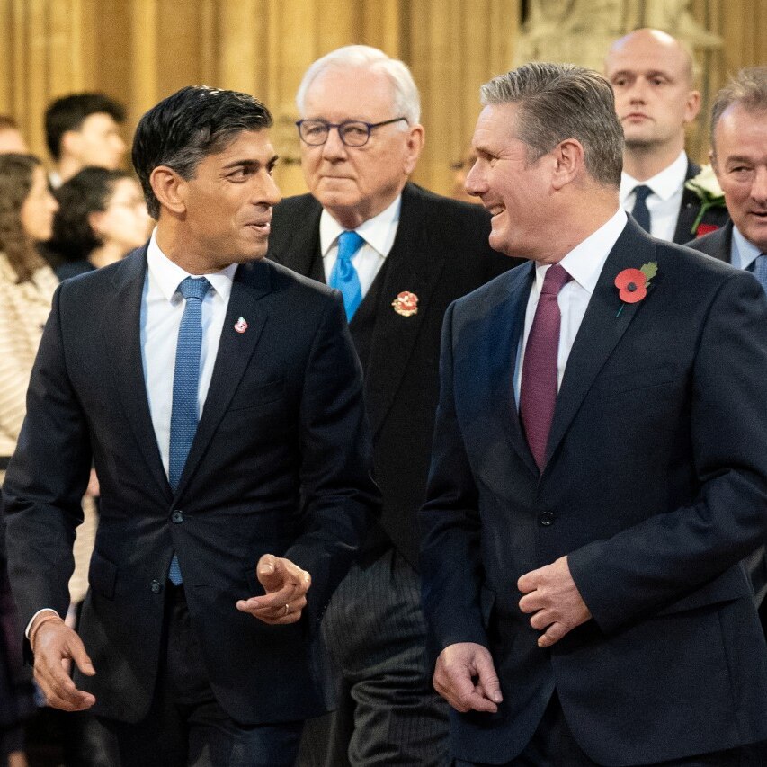 Two men in suits smiling at each other, with lines of other men behind them.,