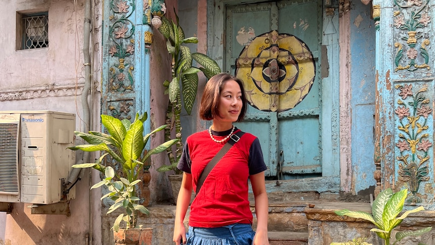 Write Maggie Zhou stands on steps in Dehli wearing a red tank over a black t-shirt and a long denim skirt.