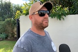 Man with  dark sunglasses and brown hat stands outside a white fence with a microphone under his chin