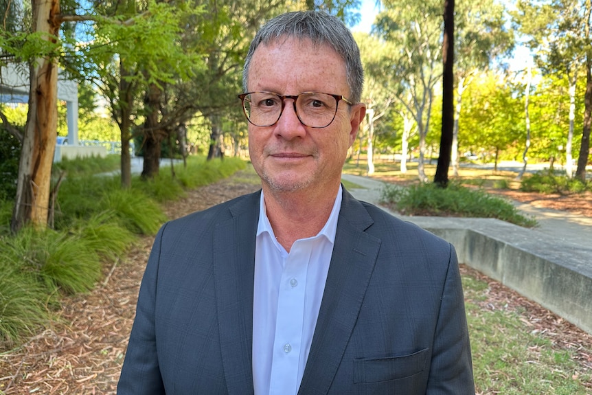 A man with greying hair and wearing a suit and glasses looks at the camera