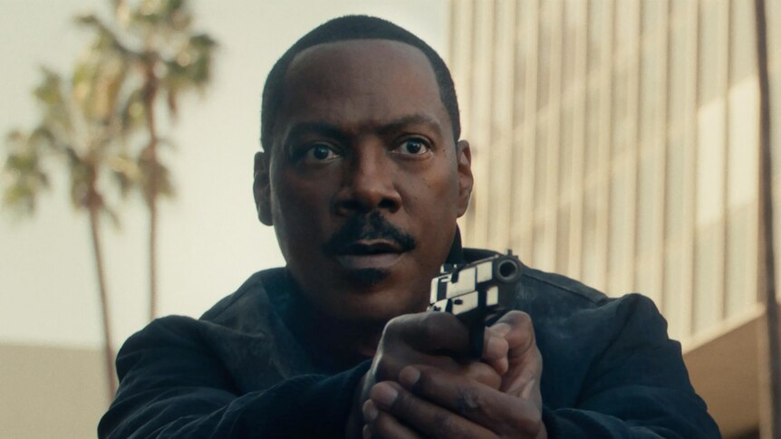 Eddie Murphy looking serious and pointing a gun with both hands, palm trees behind him