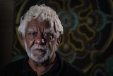 A phot of an Indigenous man looking directly at the camera with Indigenous mural in background.