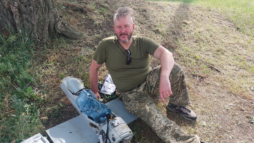 A man in a khaki Tshirt and camouflage reclines on a piece of military equipment