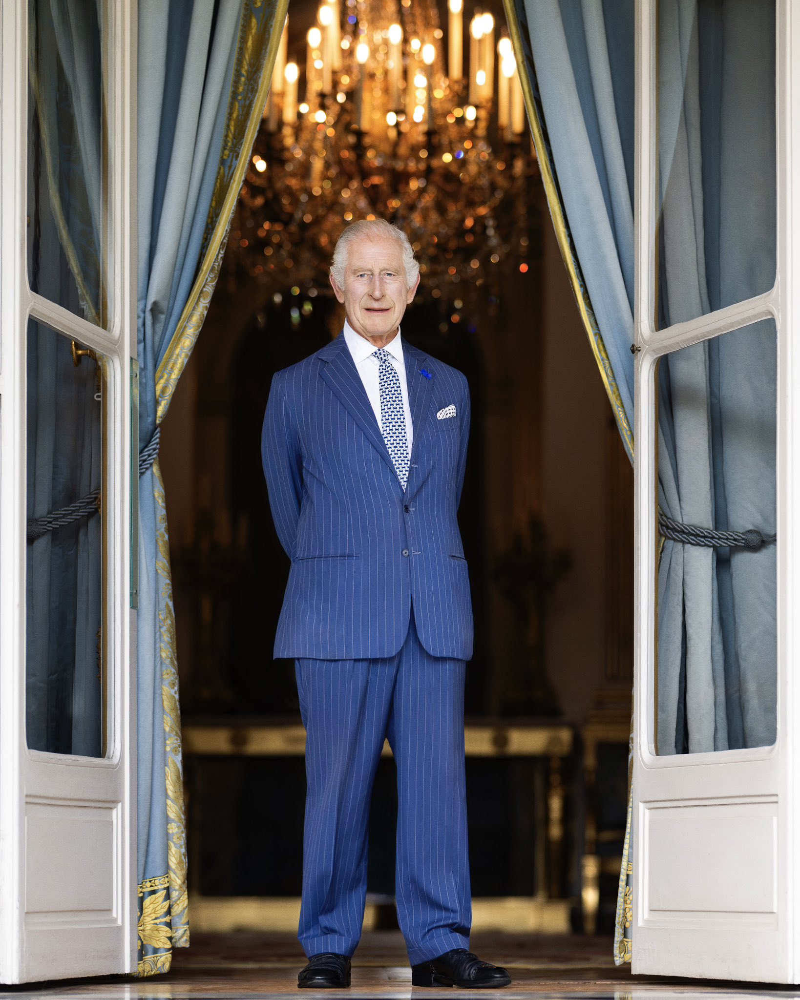 King Charles stands with his hands behind his back in an open window.