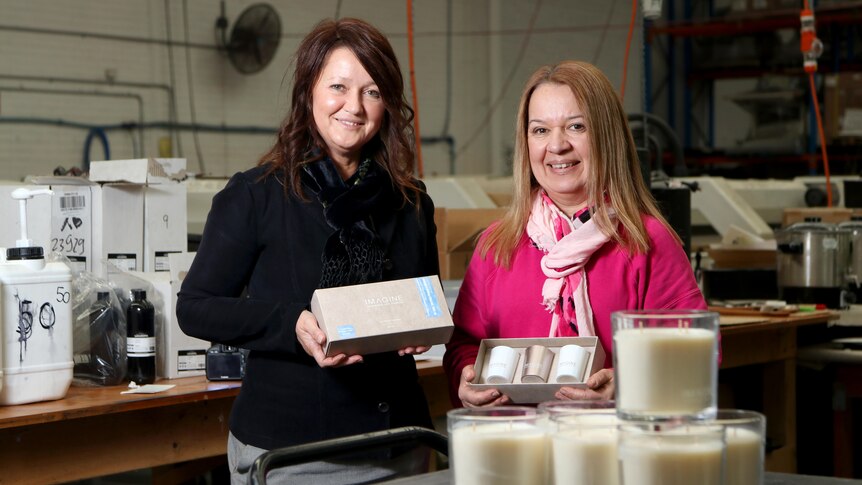 Two smiling women hold boxes containing candles while they stand in a warehouse.