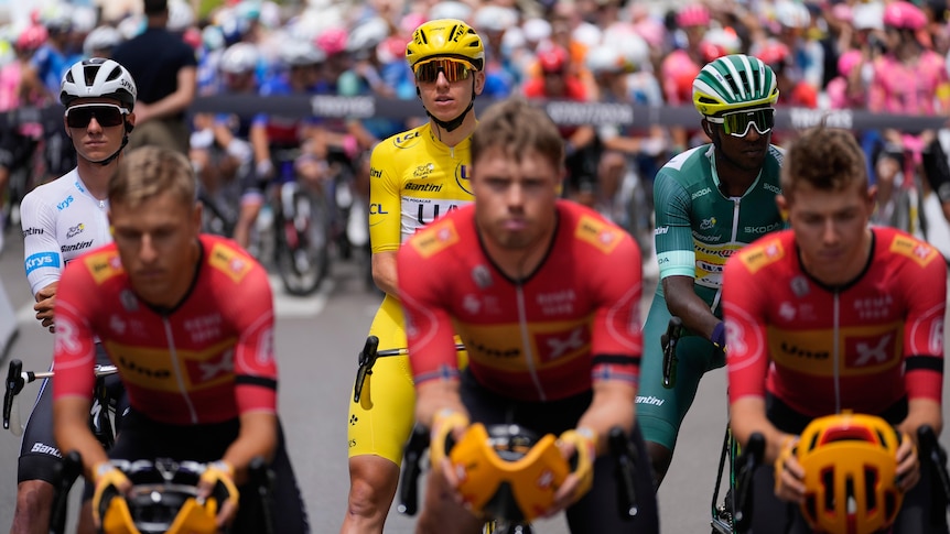 A group of cyclists in red stare silently at the camera, with the race leaders lined up behind them.